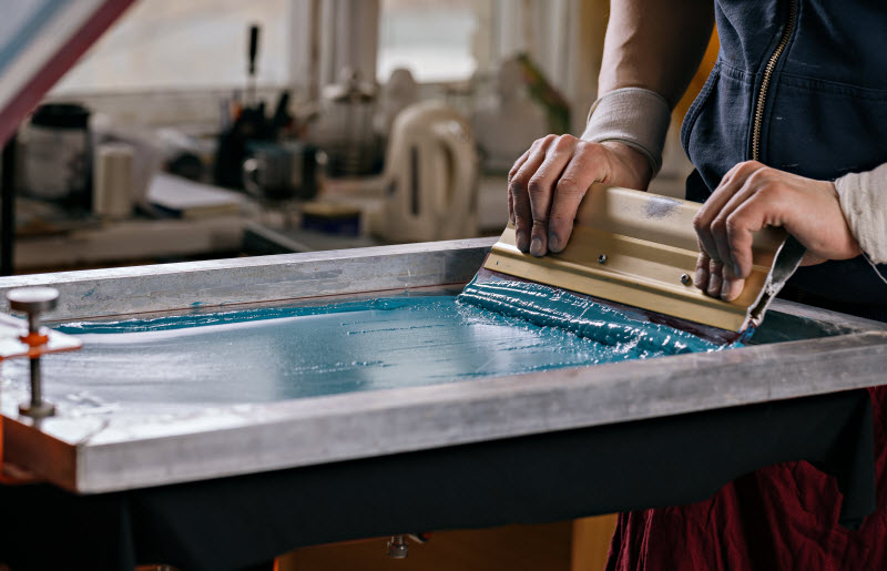 male hands with squeegee printing images on t-shirts by silkscreen method in a design studio
