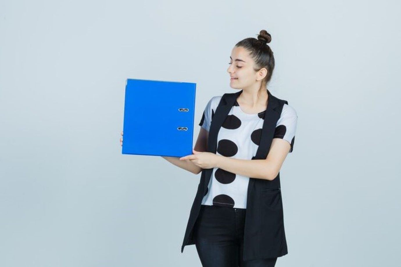 Woman in a black vest and polka-dot shirt holds a blue binder, presenting it to the side.